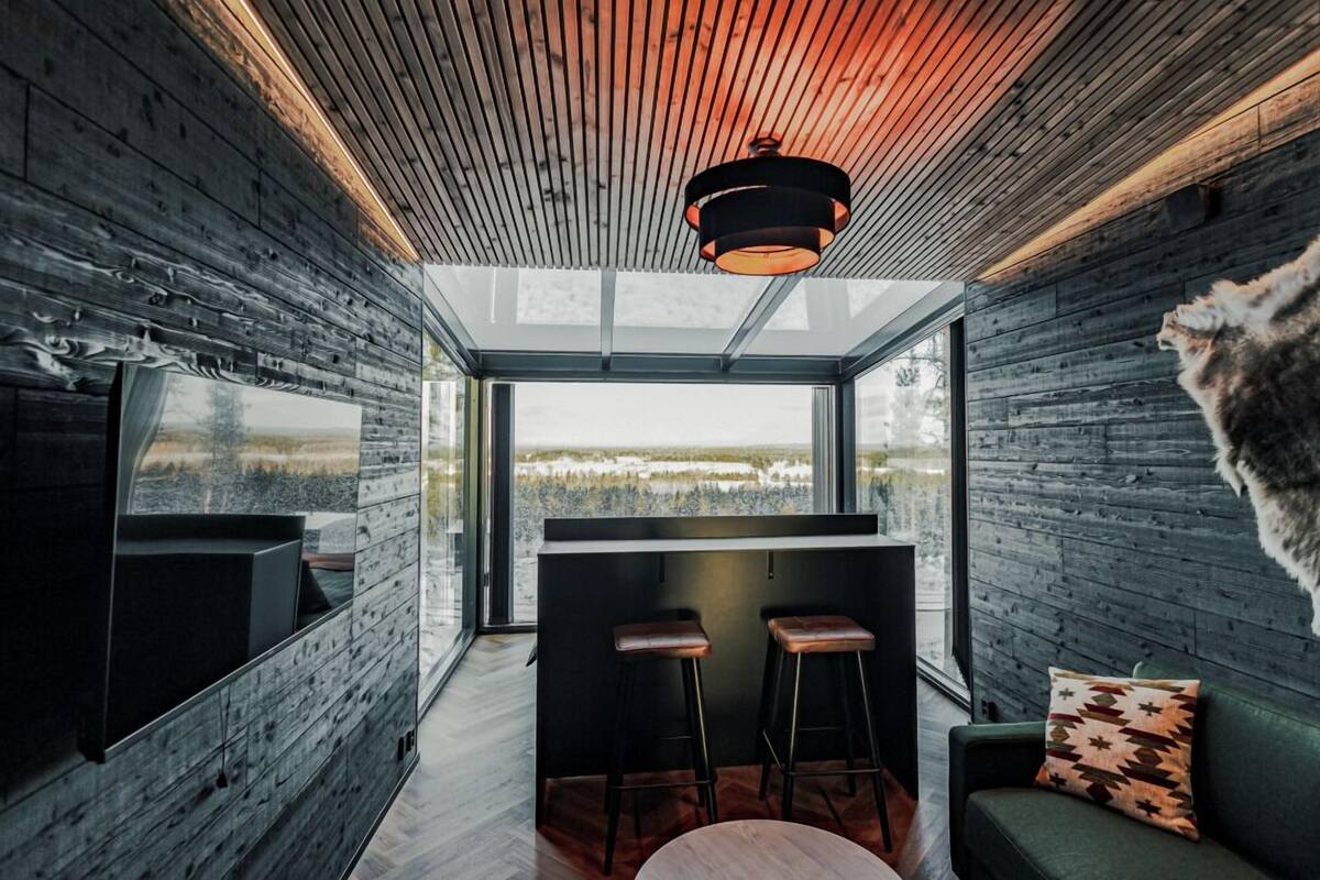 A modern interior with a wooden ceiling, a large window offering a view of a snowy landscape, a bar area with stools, and a green sofa with a patterned pillow.
