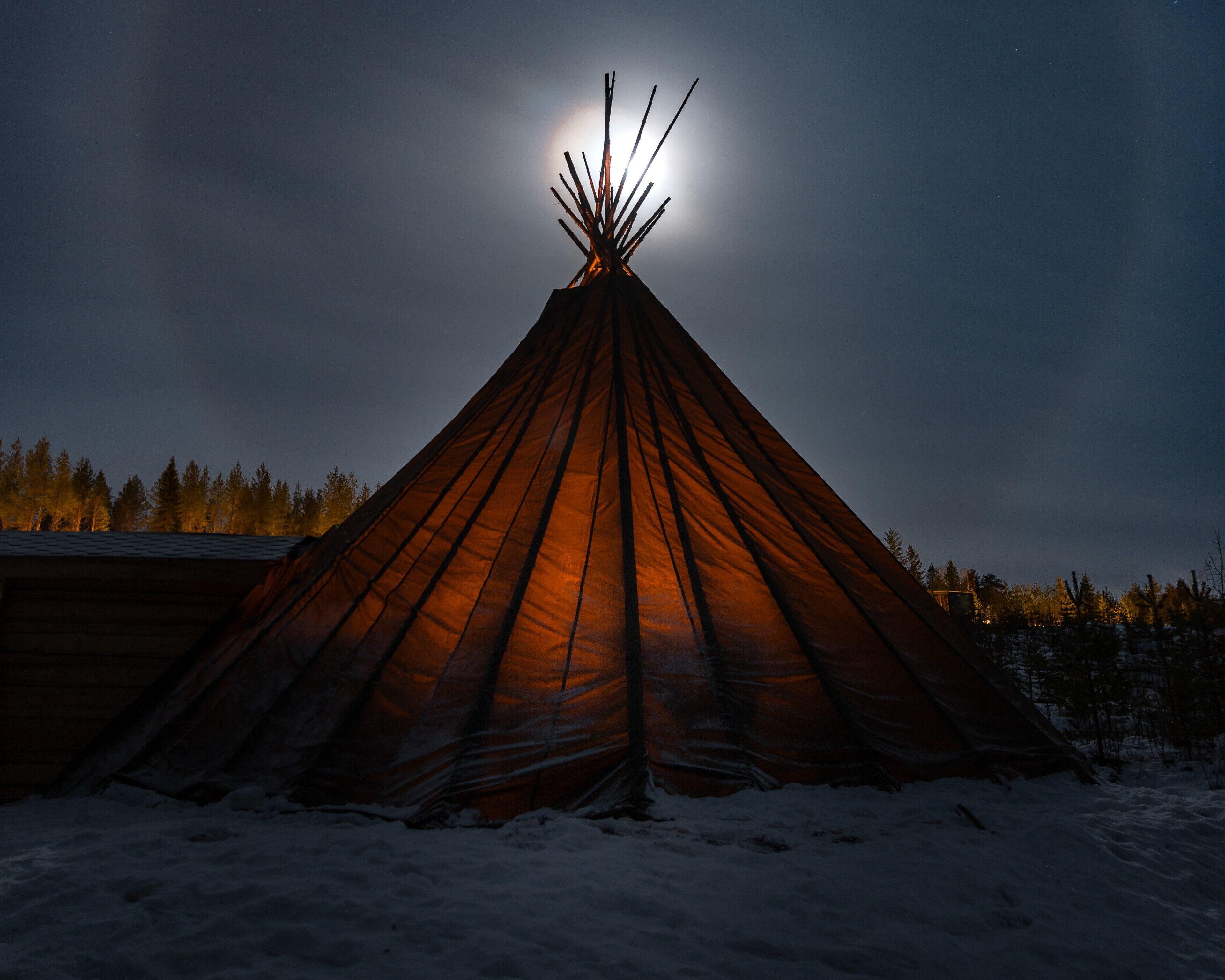 Tipi coberto de neve com uma fogueira no topo, rodeado por árvores e um edifício ao fundo.