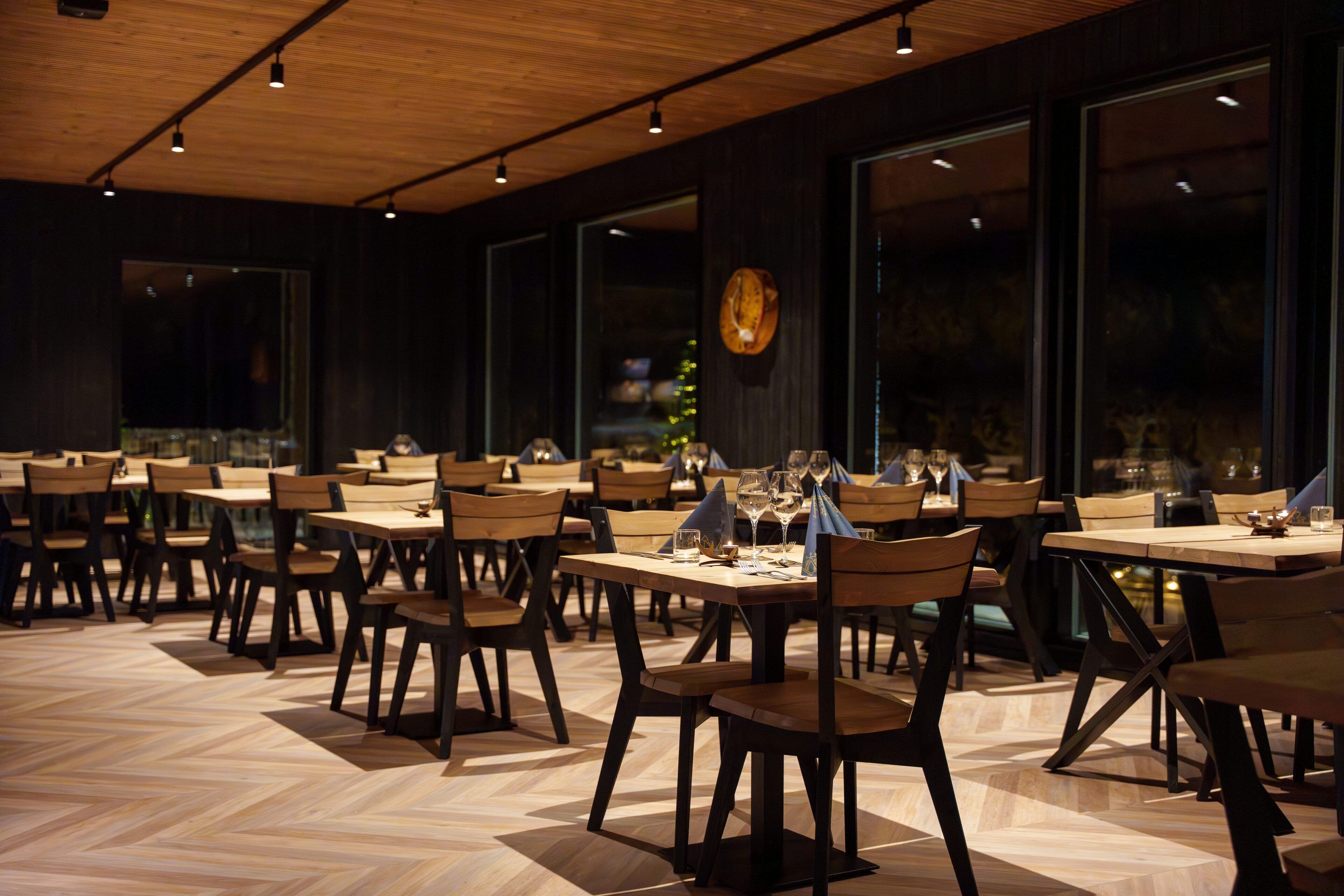 Un restaurant avec des tables et des chaises en bois, de grandes fenêtres et un parquet disposé en motif chevrons.
