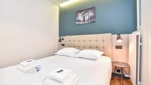 A neatly made bed with white linens and pillows, a small round table with a lamp, and a framed picture on the wall. - Authentic Apartment - 2br/8p - Near Opera Garnier (Paris)