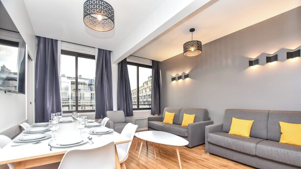 A modern living room with a grey sofa, a dining table set with white chairs, and a large window with curtains. - Authentic Apartment - 2br/8p - Near Opera Garnier (Paris)
