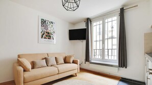 A modern living room with a beige sofa, a round rug, a flat-screen TV mounted on the wall, and a large window with curtains. - Cosy Apartment - 1br/4p - Place de la Nation (Paris)