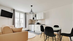 A modern living room with a sofa, dining table, and chairs. A kitchen area is visible in the background with white cabinets and a flat-screen TV mounted on the wall. - Cosy Apartment - 1br/4p - Place de la Nation (Paris)