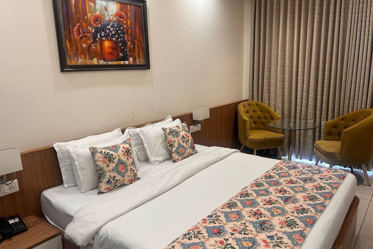 A hotel room with a bed, two floral-patterned pillows, a bedside table with a phone, a painting on the wall, and a window with curtains.