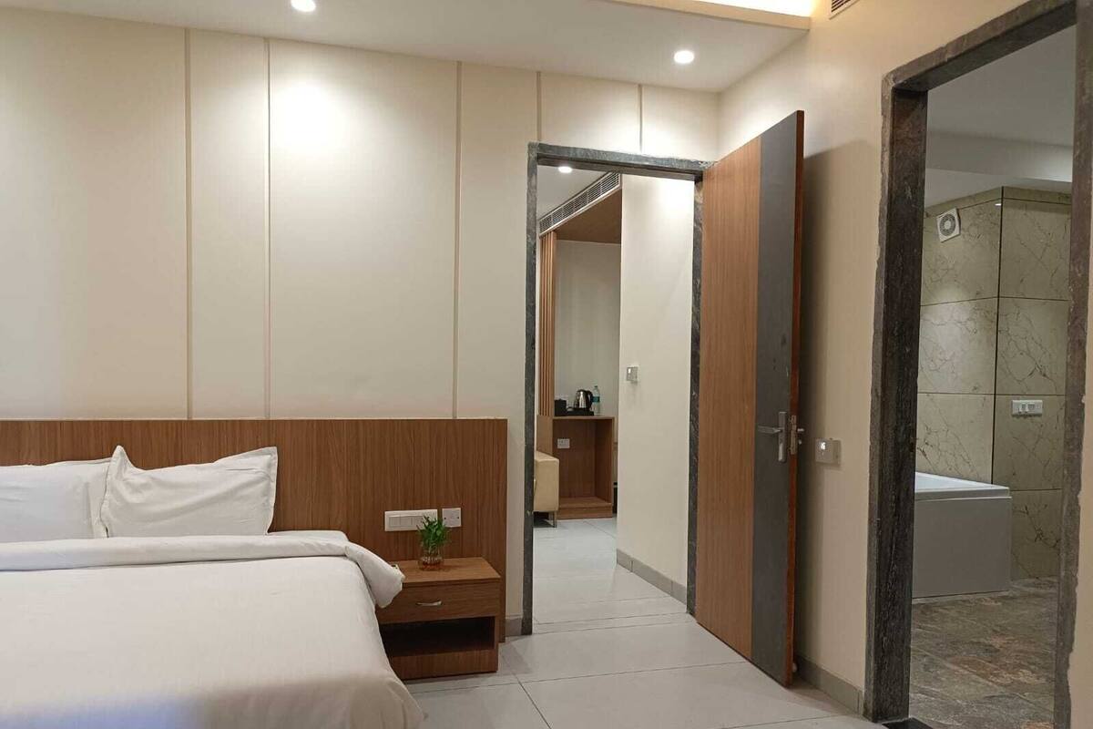 A modern hotel room with a large bed, a wooden nightstand, a small plant, and a bathroom visible through an open door.