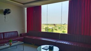 A modern living room with a red sofa, a glass coffee table, and a view of a green landscape through large windows with red curtains. - THE PRINCE ZENQ XPRESS RESORTT ROHTAK (Sampla)