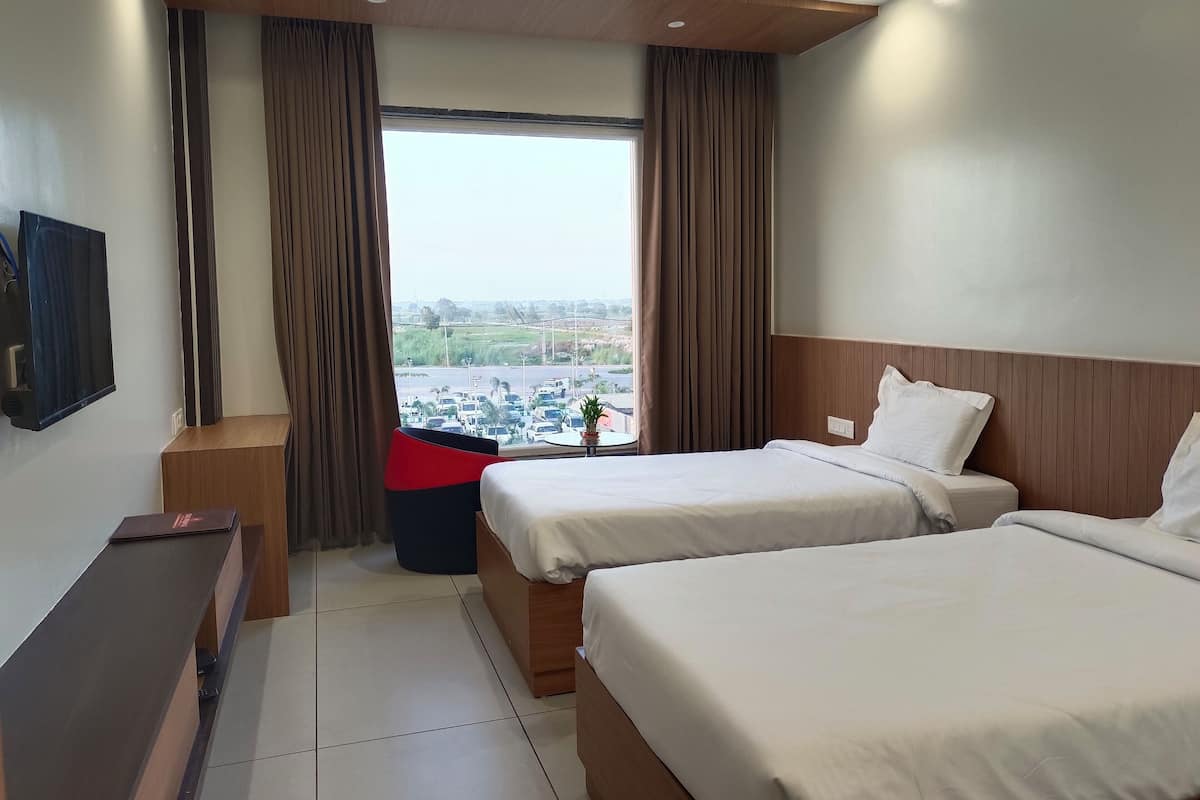 A hotel room with two beds, a TV, a desk, and a window with a view of a parking lot and buildings.