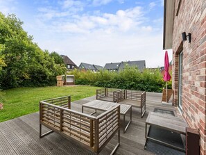 Une terrasse en bois avec des meubles d’extérieur, un bâtiment en briques et une zone herbeuse avec des arbres.