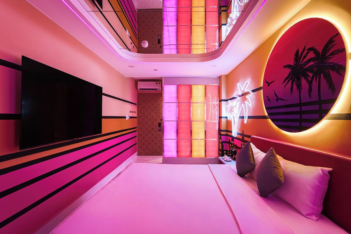 Een moderne hotelkamer met een grote flatscreen-tv, een bed met kussens, een muur met een rond neonontwerp en uitzicht op palmbomen.