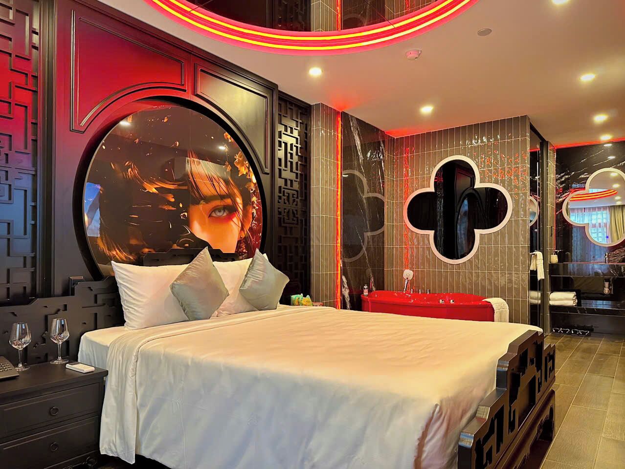 A modern hotel room with a large round mirror, a bed with white linens, a red accent wall, and a small seating area with a red sofa.
