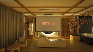 A modern room with a large screen displaying "NETFLIX," a sofa, a lounge chair, and a wooden floor.