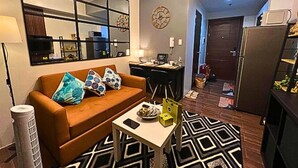 A compact living space with a sofa, a small table, a patterned rug, a refrigerator, a microwave, and a kitchen area with a counter and cabinets. - Rent Xpress Online Heart of Makati (Makati)