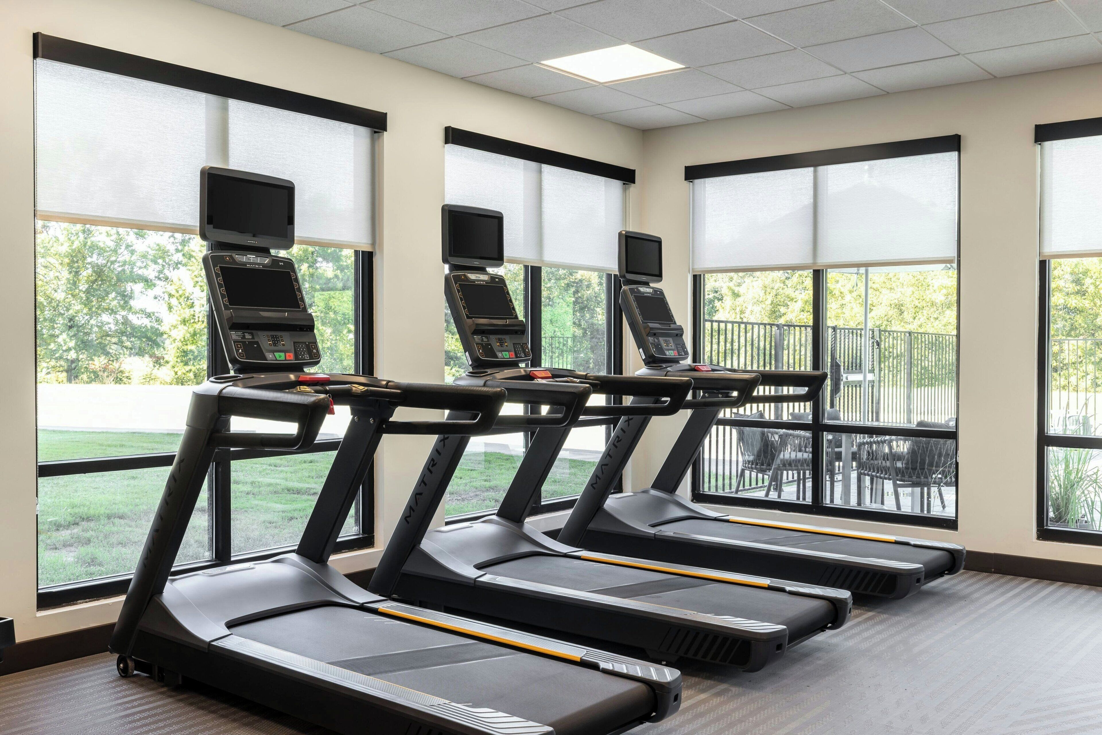 A gym with treadmills, exercise screens, and large windows with a view of greenery.