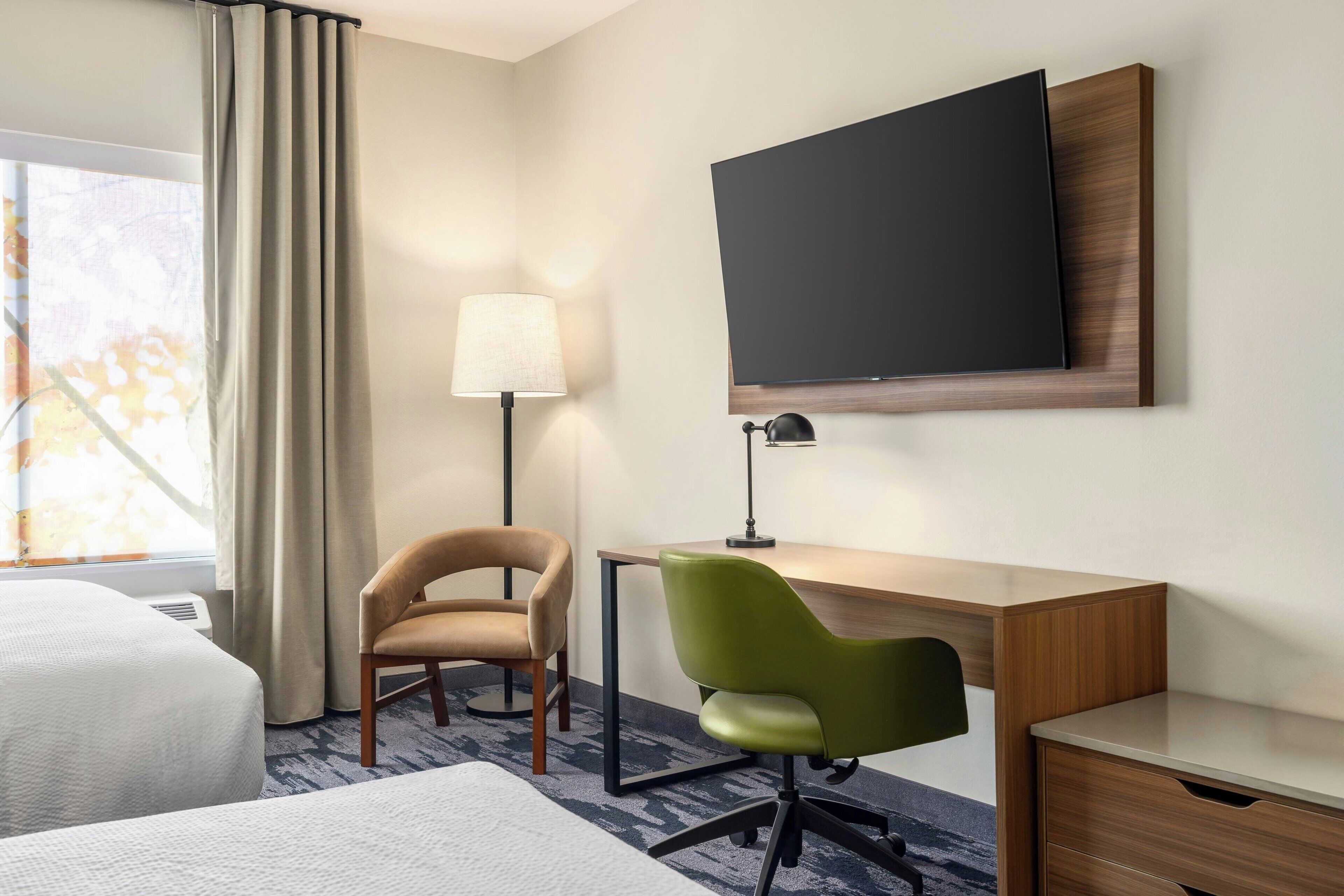 A hotel room with a bed, a desk, two chairs, a TV, and a lamp.
