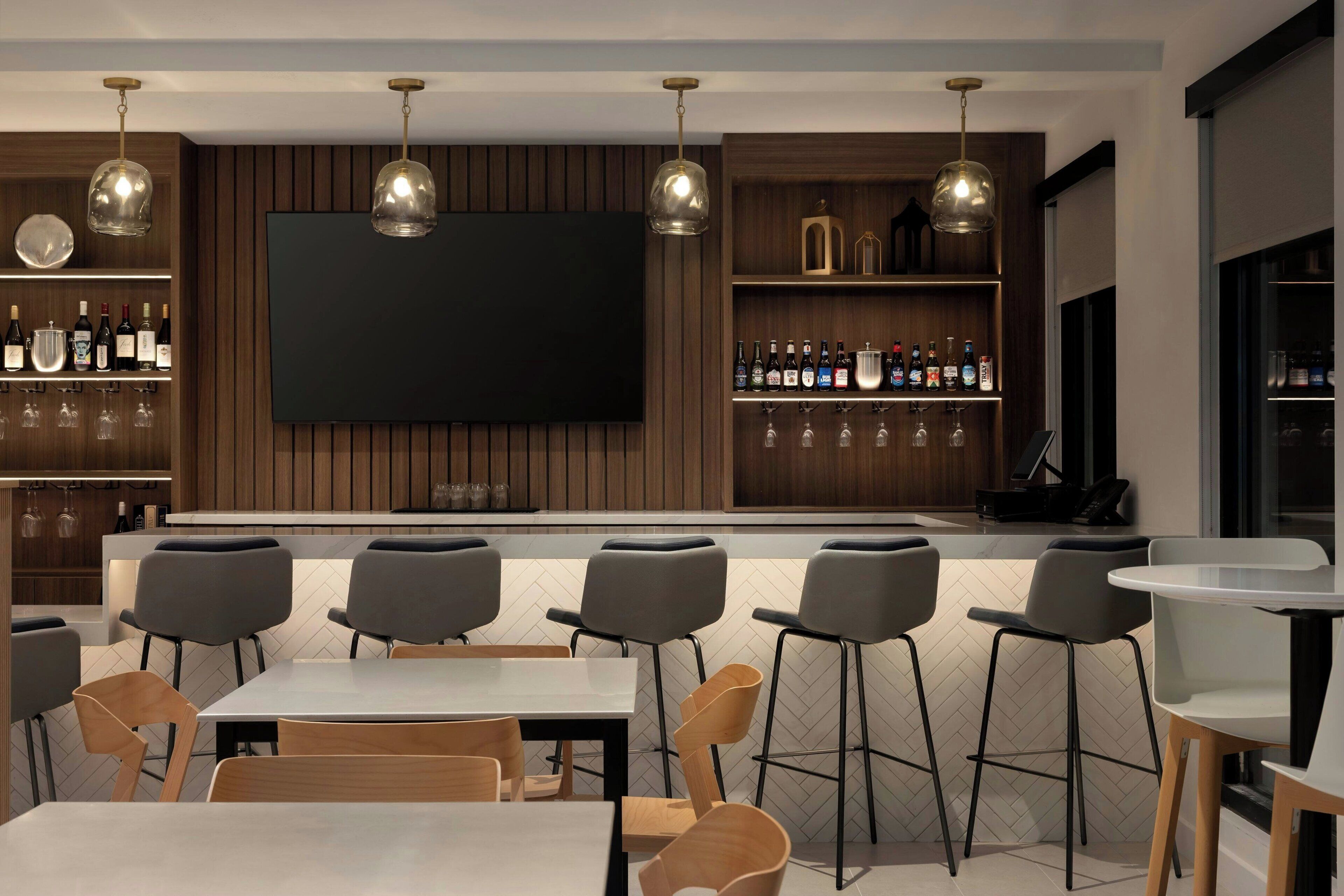A modern bar area with a flat-screen TV, wooden shelves with bottles, and bar stools.
