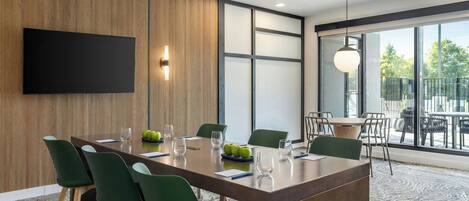 A modern conference room with a large table, green chairs, a flat-screen TV, and a large window offering a view of the outdoors.
