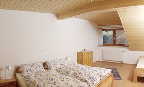 Ett sovrum med ett sluttande tak, en säng med blommigt sänglinne, ett nattduksbord med en lampa, ett fönster med gardiner och ett element.