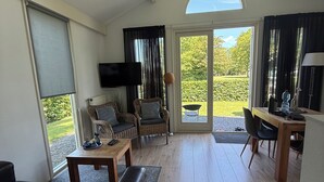 A spacious living area with a sofa, a dining table, and a TV. There is a large window providing a view of the outdoors. - Cozy vacation home in North Holland in a very quiet park, pure relaxation! (Dirkshorn)