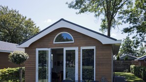 A small, single-story building with a sloped roof, a large glass door, and a small patio area with outdoor seating. - Cozy vacation home in North Holland in a very quiet park, pure relaxation! (Dirkshorn)