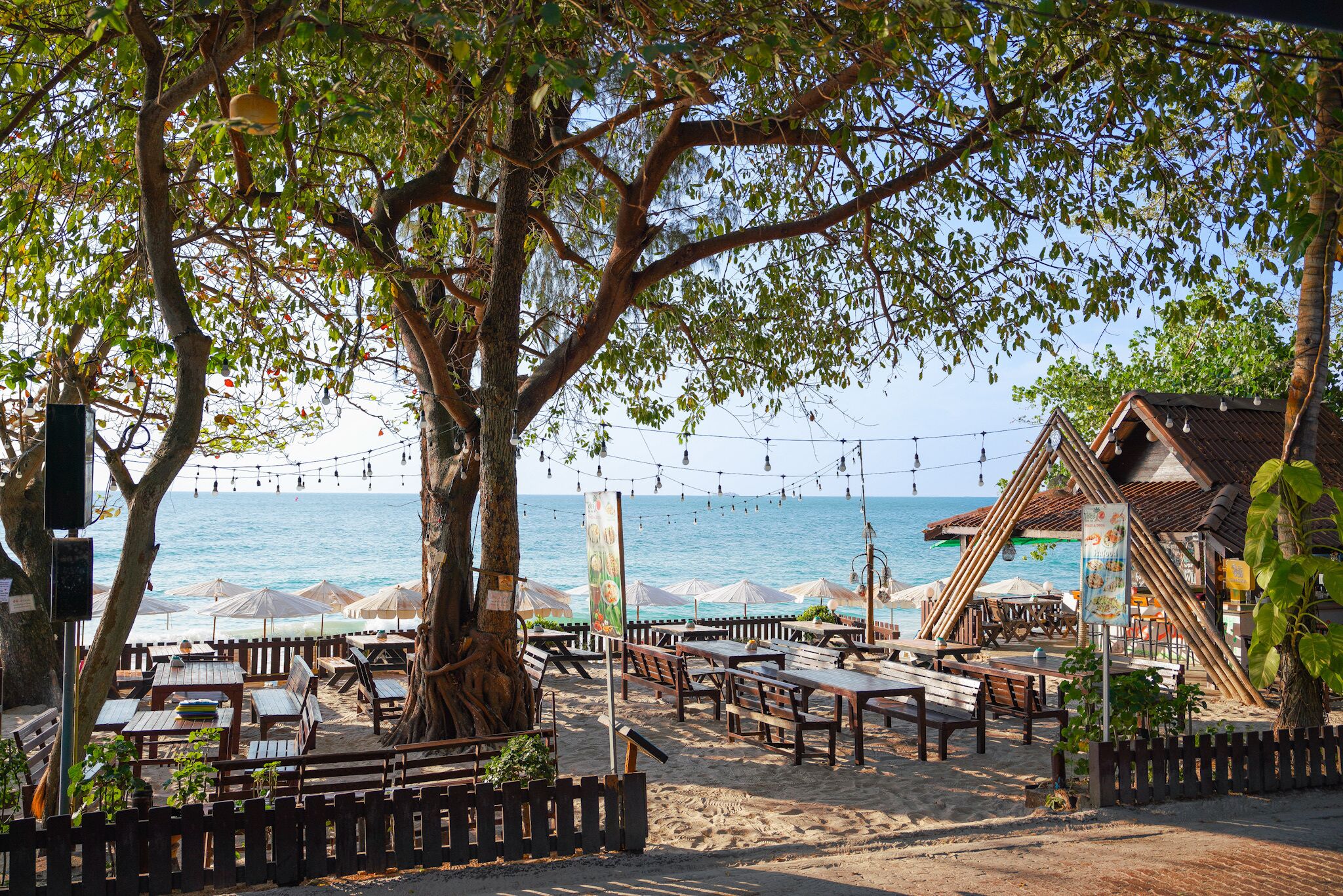 Un restaurante frente a la playa con terraza, valla de madera y luces colgantes.