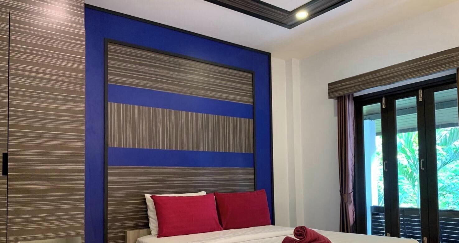 A bedroom with a bed, a nightstand, a small refrigerator, and a window with a view of greenery.