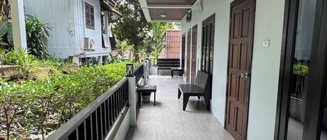 Un pasillo exterior cubierto con un banco de madera y una mesa, rodeado de vegetación y un edificio blanco con puertas oscuras.