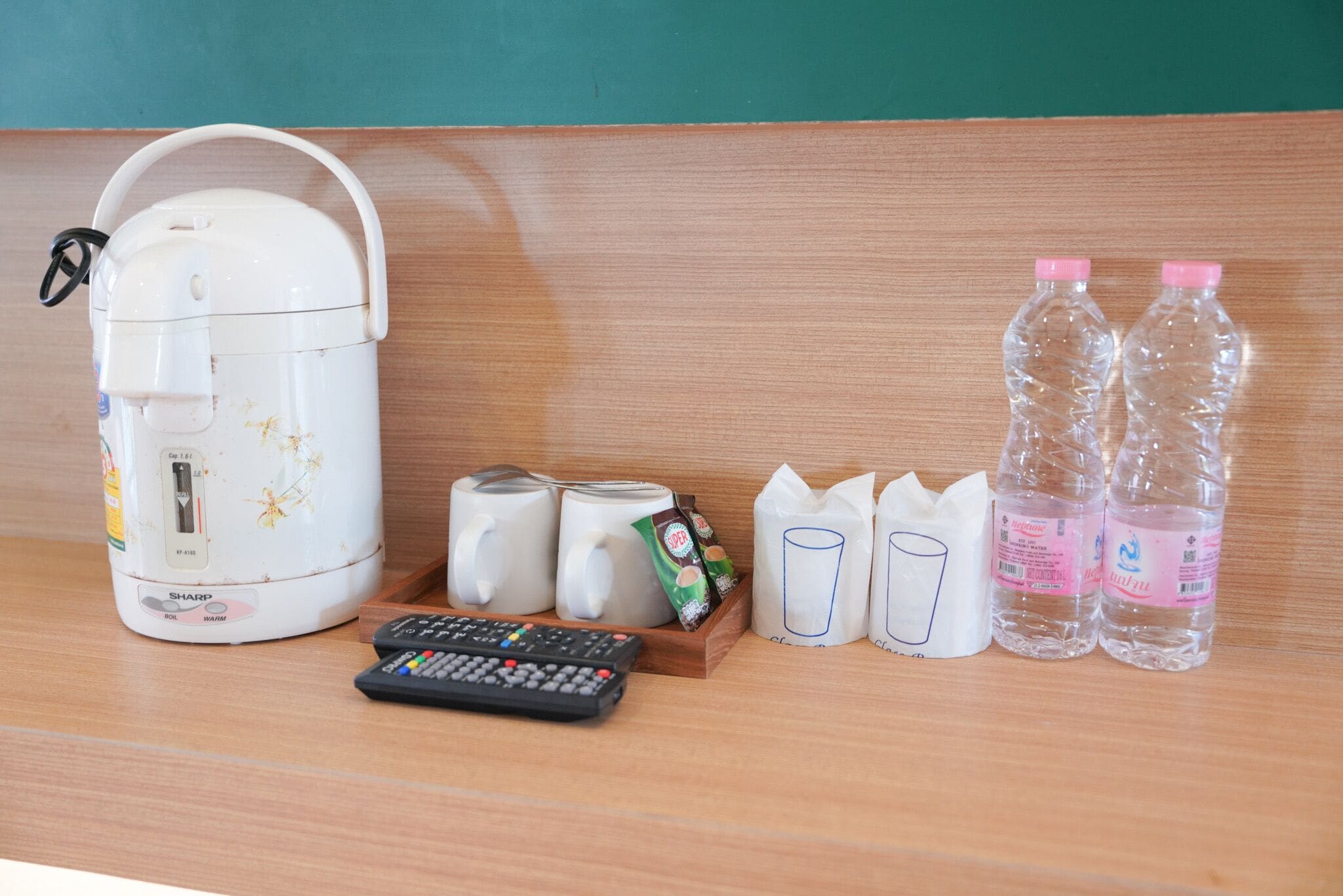 Un área de cocina con una cocedora de arroz, dos tazas, un control remoto, un paquete de fideos instantáneos, dos botellas de agua y vasos de papel.