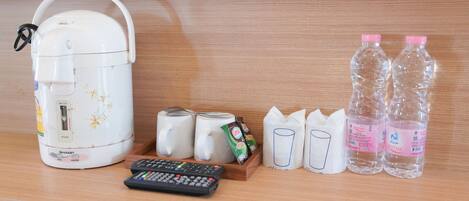 A kitchen area with a rice cooker, two mugs, a remote control, a packet of instant noodles, two water bottles, and paper cups.