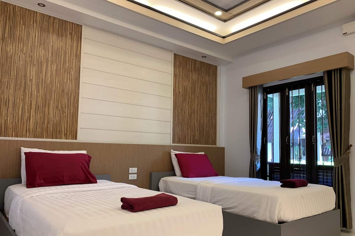 A modern hotel room with two beds, each with a wooden headboard and red pillows. The room has a large window with curtains, a ceiling light, and a wooden wall panel.