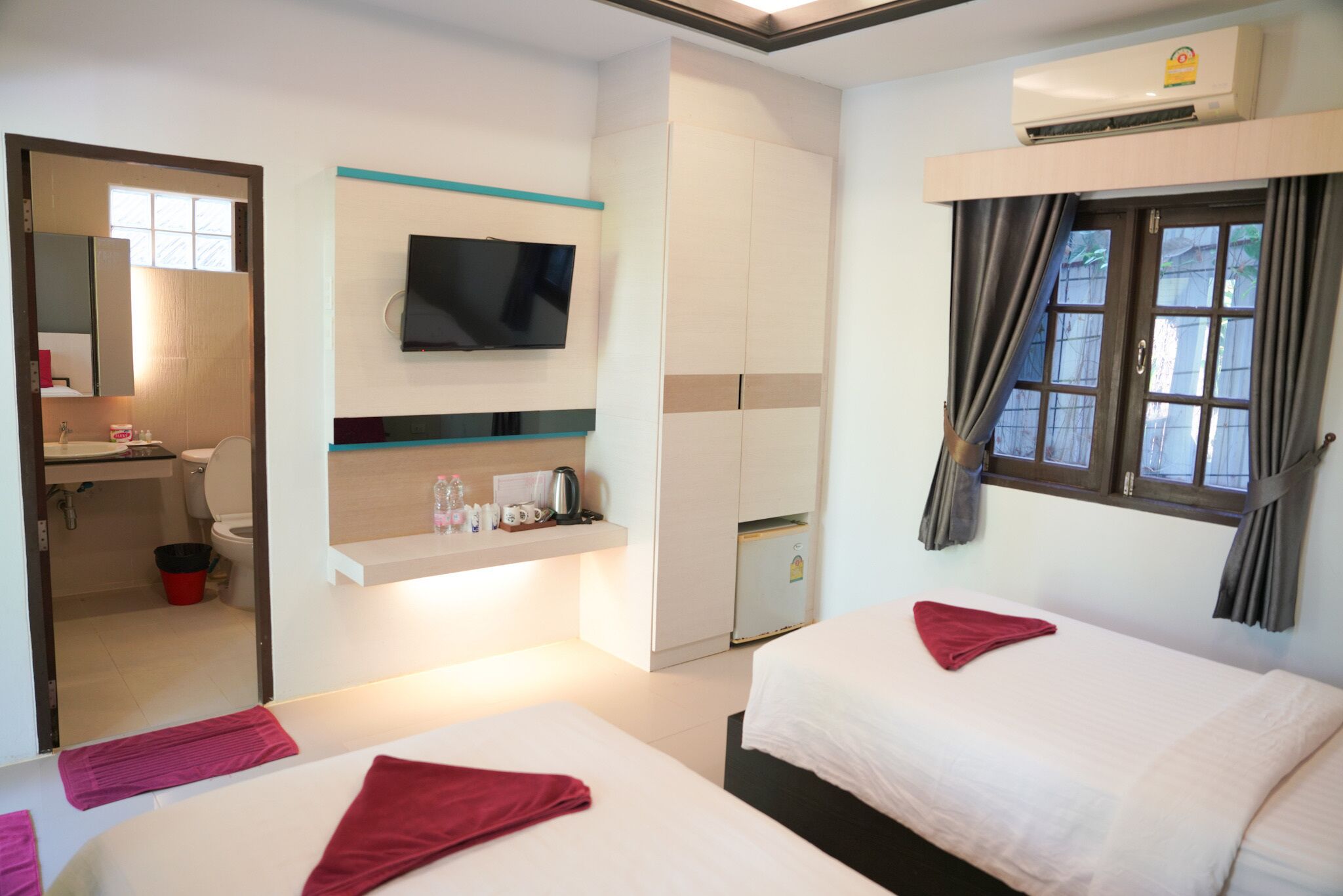 Habitación de hotel con dos camas, un televisor montado en la pared, una kitchenette pequeña y una ventana con cortinas.