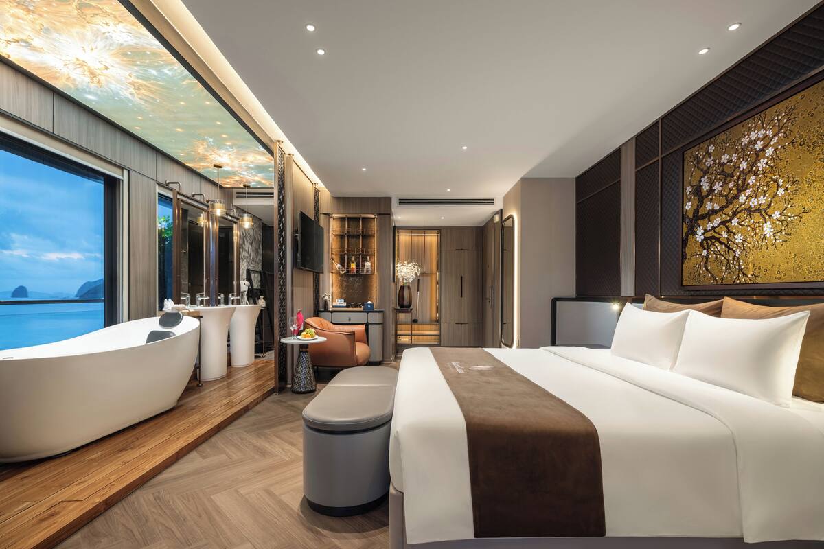 A modern hotel room with a large bed, a bathtub, a vanity, and a large window overlooking the sea.