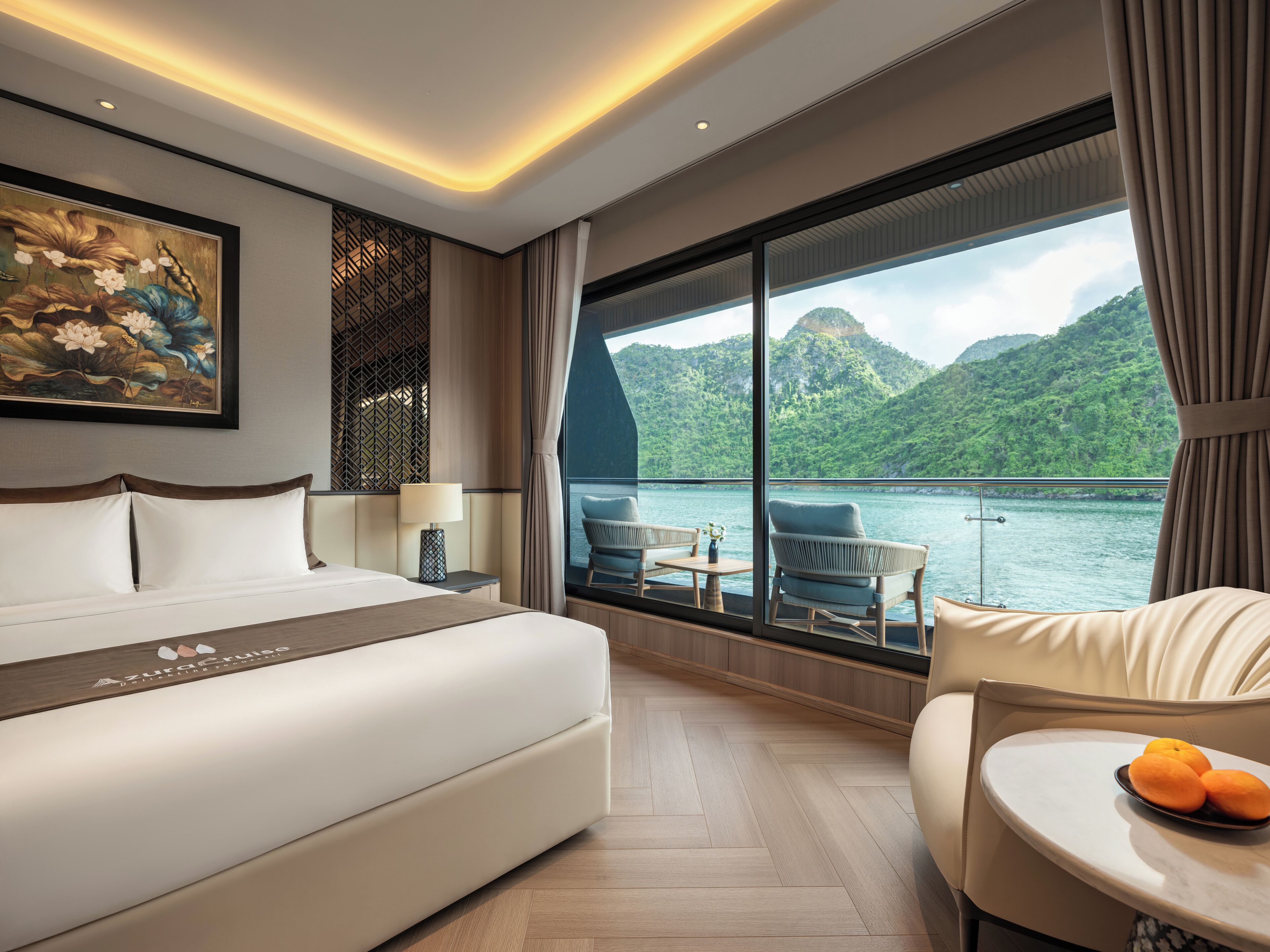 Habitación de hotel con cama, zona de estar con mesa, ventana grande con vista a un lago y montañas, y un cuadro en la pared.