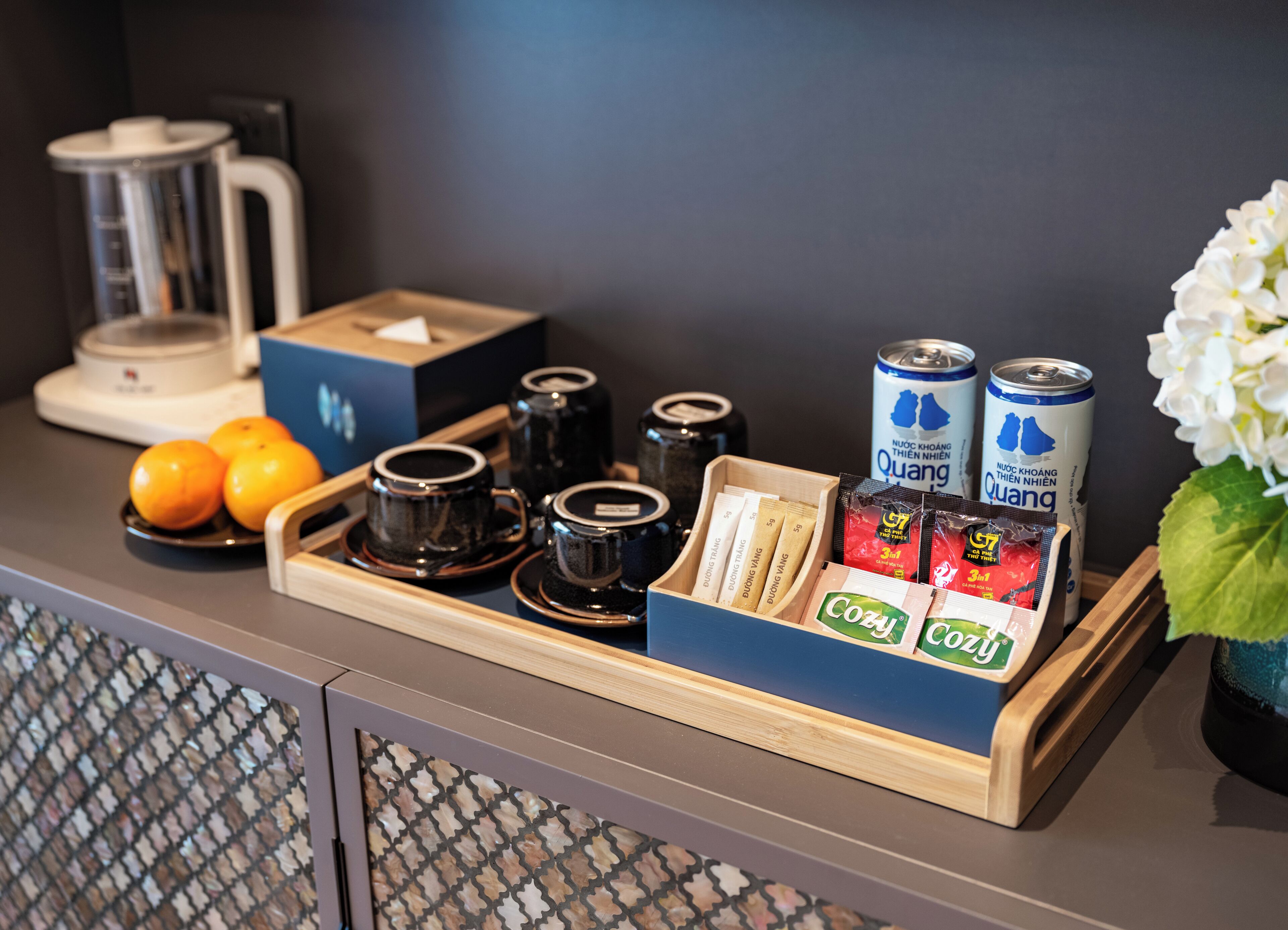 A tray with coffee cups, oranges, tea bags, and cans of Quang beverage on a hotel counter.