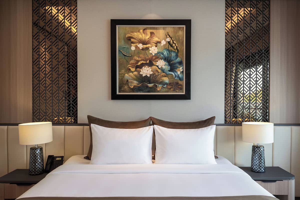 A hotel room with a bed, two bedside tables with lamps, a framed artwork above the bed, and a decorative wall paneling.