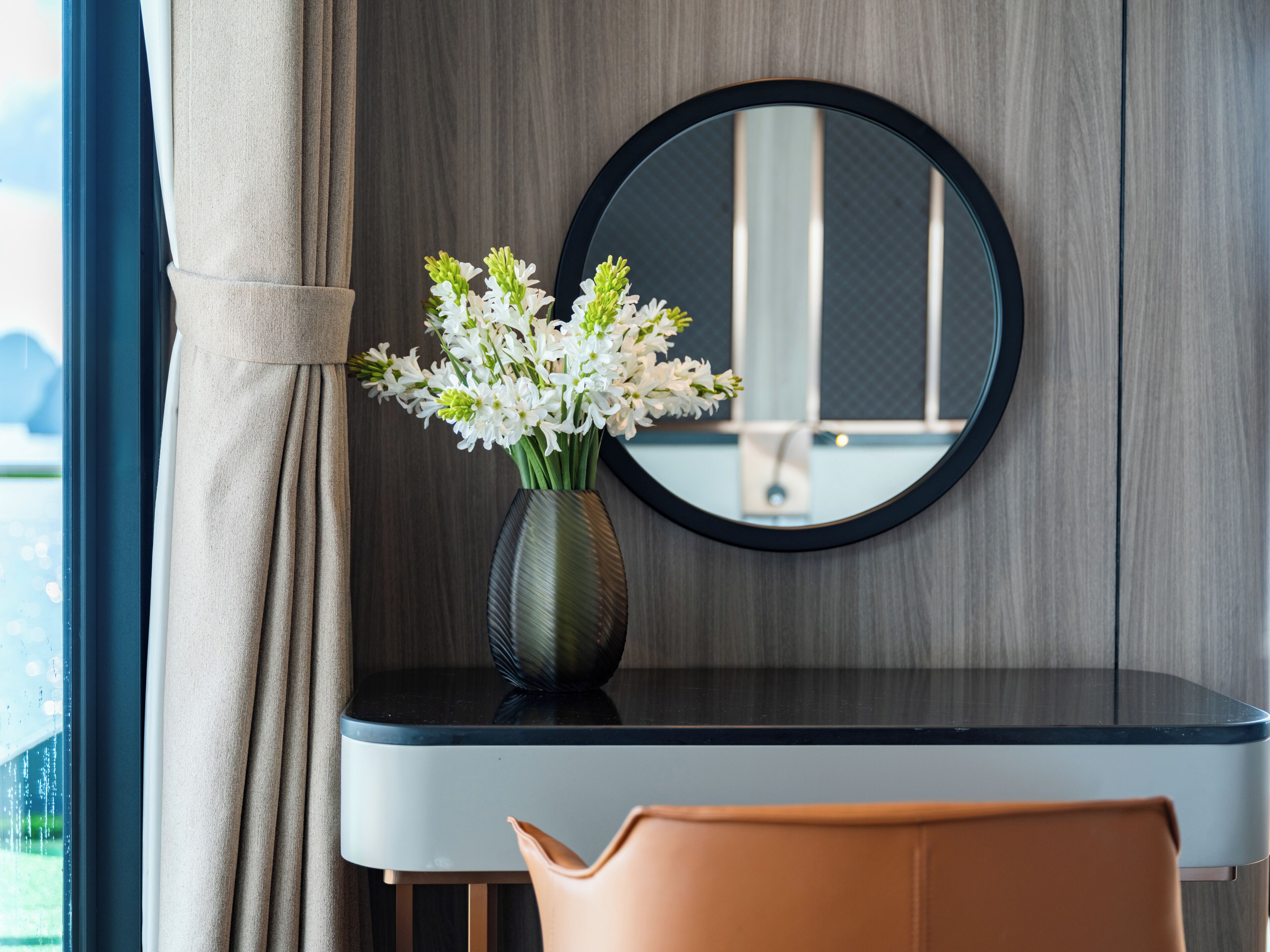 A room with a round mirror, a vase with flowers, a table, and a chair.
