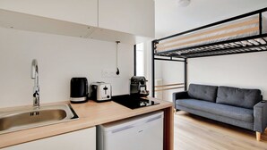 Eine kompakte Küchenzeile mit Spüle, Toaster und Kaffeemaschine, ein graues Sofa und ein Etagenbett.