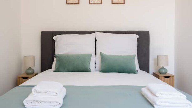 Ein ordentlich gemachtes Bett mit weißen und grünen Kissen, zwei Nachttische mit Lampen und einer hellblauen Tagesdecke.