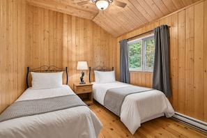 A room with two single beds, wooden walls, a window with curtains, and a ceiling fan.