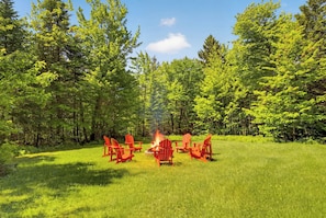 A campsite with a fire pit surrounded by red chairs in a grassy field with dense trees in the background.