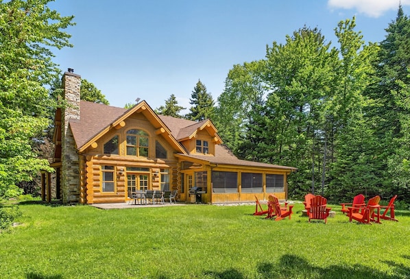 A log cabin with a large porch, outdoor seating, and a view of the surrounding forest.