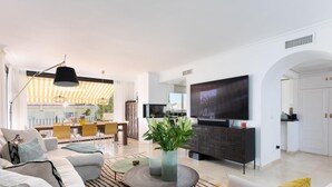 Una sala de estar moderna con un sofá, una mesa de centro y un televisor. Al fondo, se distingue un comedor con sillas amarillas y un mantel a rayas.