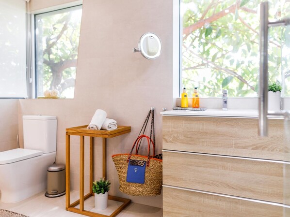 A bathroom with a toilet, a sink, a mirror, a small table with towels, a trash can, a basket with a bag, and toiletries on the windowsill. - Luxurious Villa La Paz Beach Port Aventura Golf (Salou)