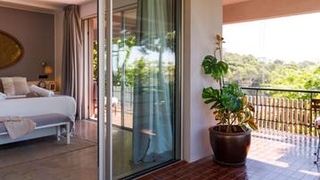 A hotel room with a bed, a potted plant, and a balcony with a view of greenery.