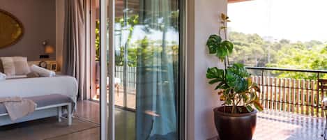 A hotel room with a bed, a potted plant, and a balcony with a view of greenery.