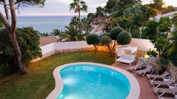 A swimming pool with a curved edge, surrounded by a stone wall and a grassy area. There are lounge chairs and a small table with a canopy.