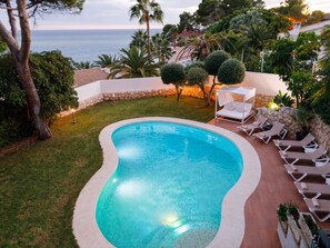 A swimming pool with a curved edge, surrounded by a stone wall and a grassy area. There are lounge chairs and a small table with a canopy. - Luxurious Villa La Paz Beach Port Aventura Golf (Salou)