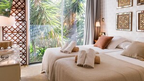 Een hotelkamer met een grote schuifglazen deur die uitkomt op een tropische tuin, een bed met keurig opgevouwen handdoeken en decoratieve muurkunst.