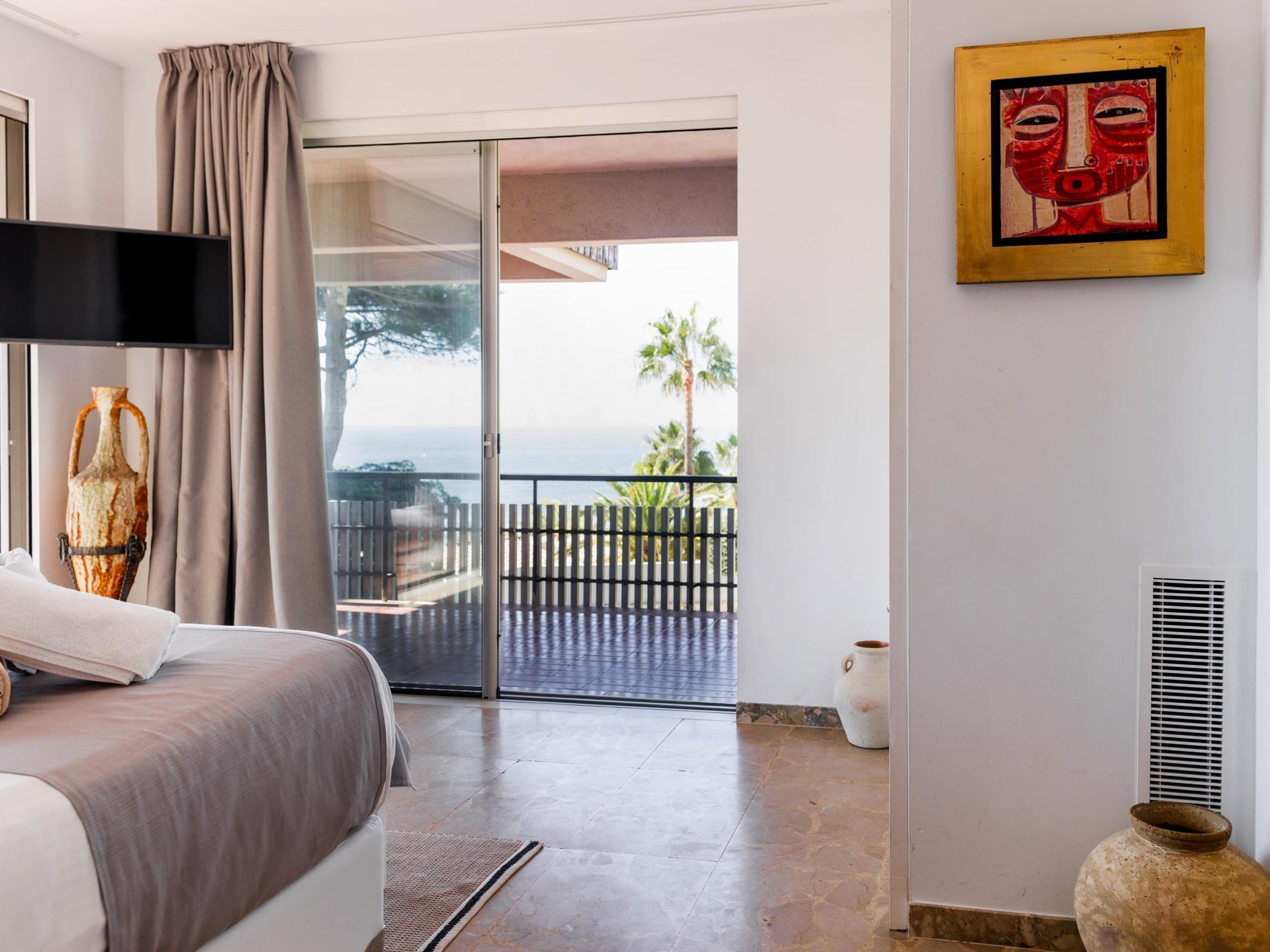 A bedroom with a bed, a flat-screen TV, a large window with curtains, a view of the sea, a balcony with a wooden fence, and a painting on the wall.