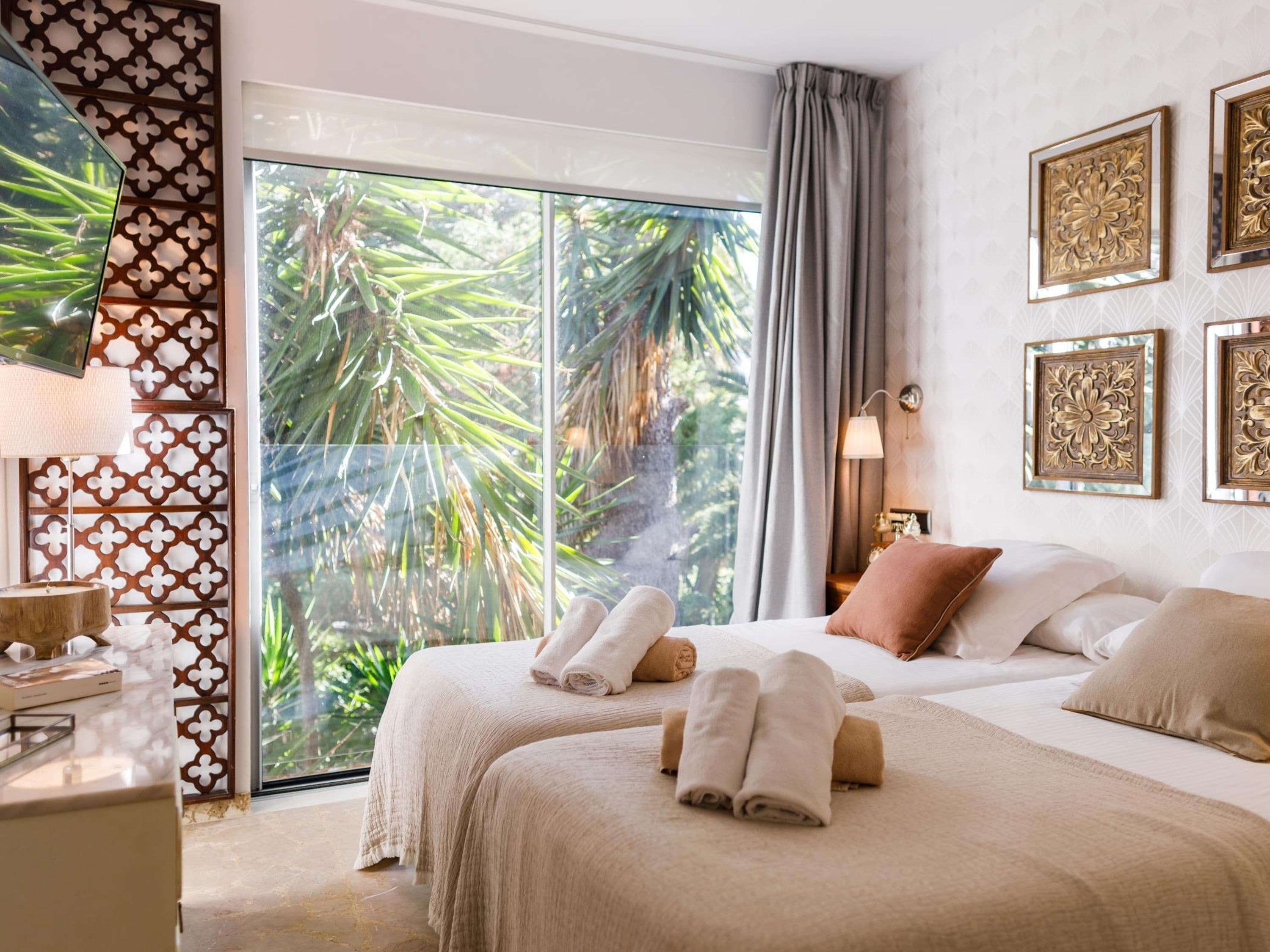 Habitación de hotel con un amplio ventanal corredero que da a un jardín tropical, una cama con toallas ordenadas y decoración en la pared.