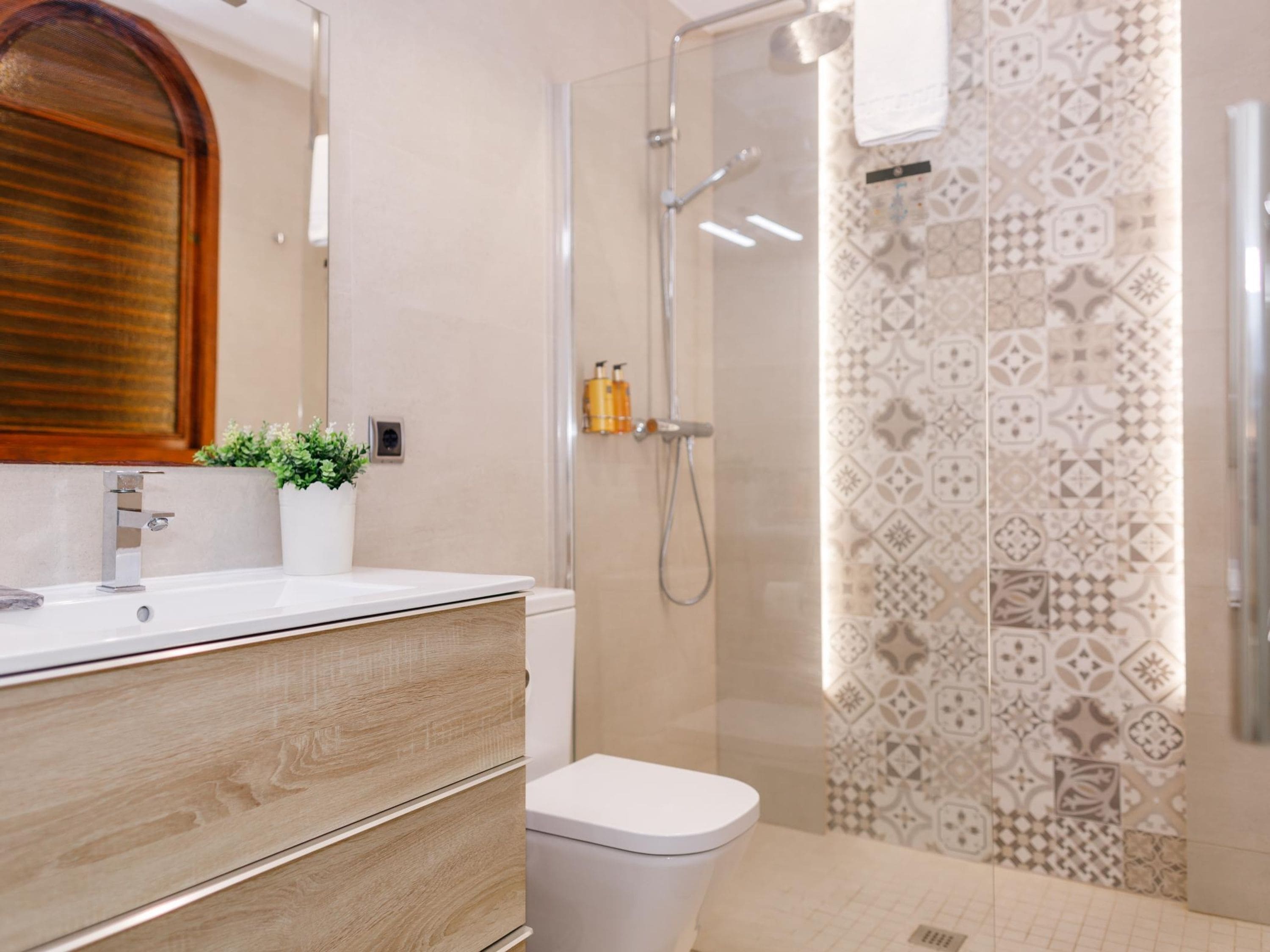 A modern bathroom with a wooden vanity, a white toilet, a glass shower enclosure, and patterned tiles.